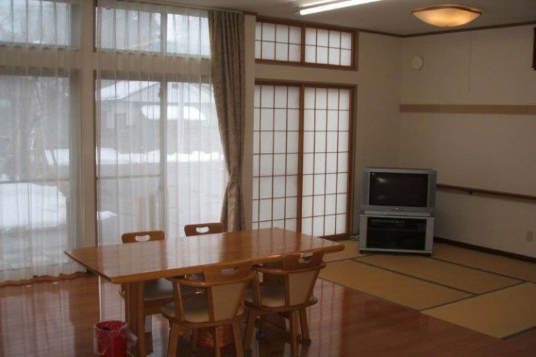 木製テーブルと畳敷きの共用スペース。大窓から自然光が入る明るい居間。